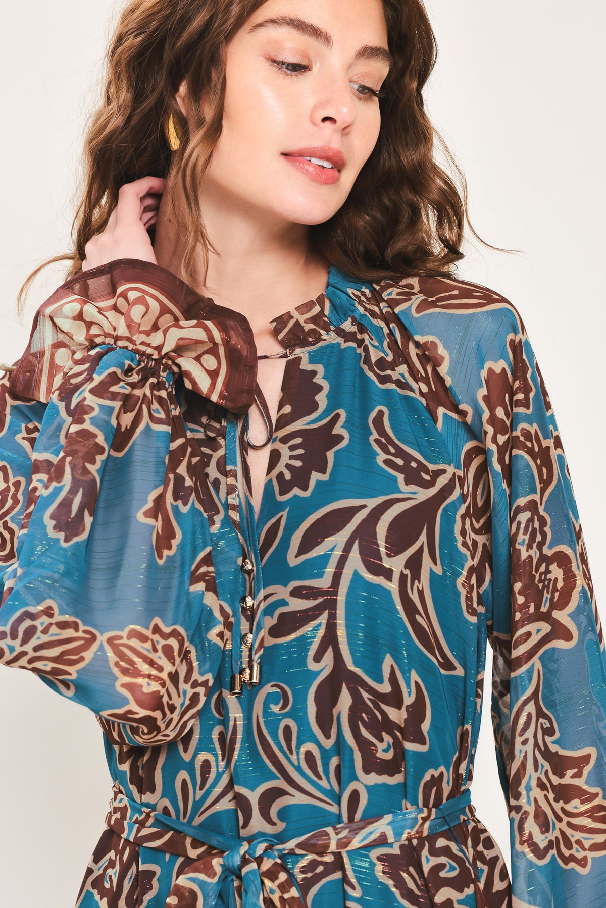 Woman wearing a teal blue long sleeve maxi dress with ruffled sleeves, tiered skirts, and brown floral print and neck tie | Flying Tomato