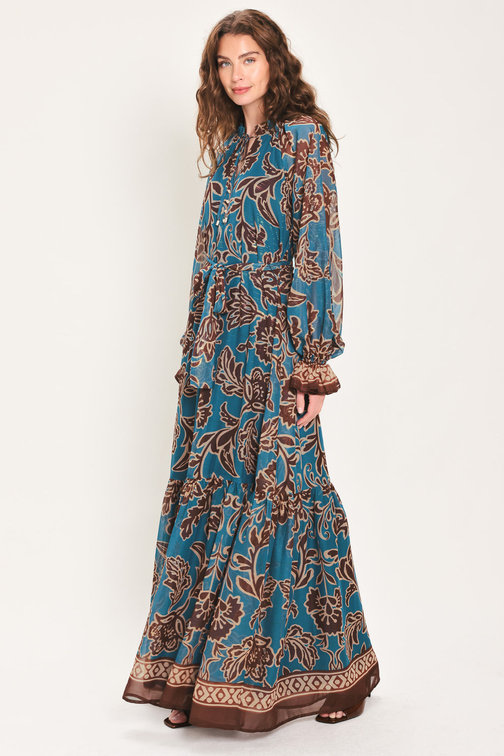 Woman wearing a teal blue long sleeve maxi dress with ruffled sleeves, tiered skirts, and brown floral print and neck tie | Flying Tomato