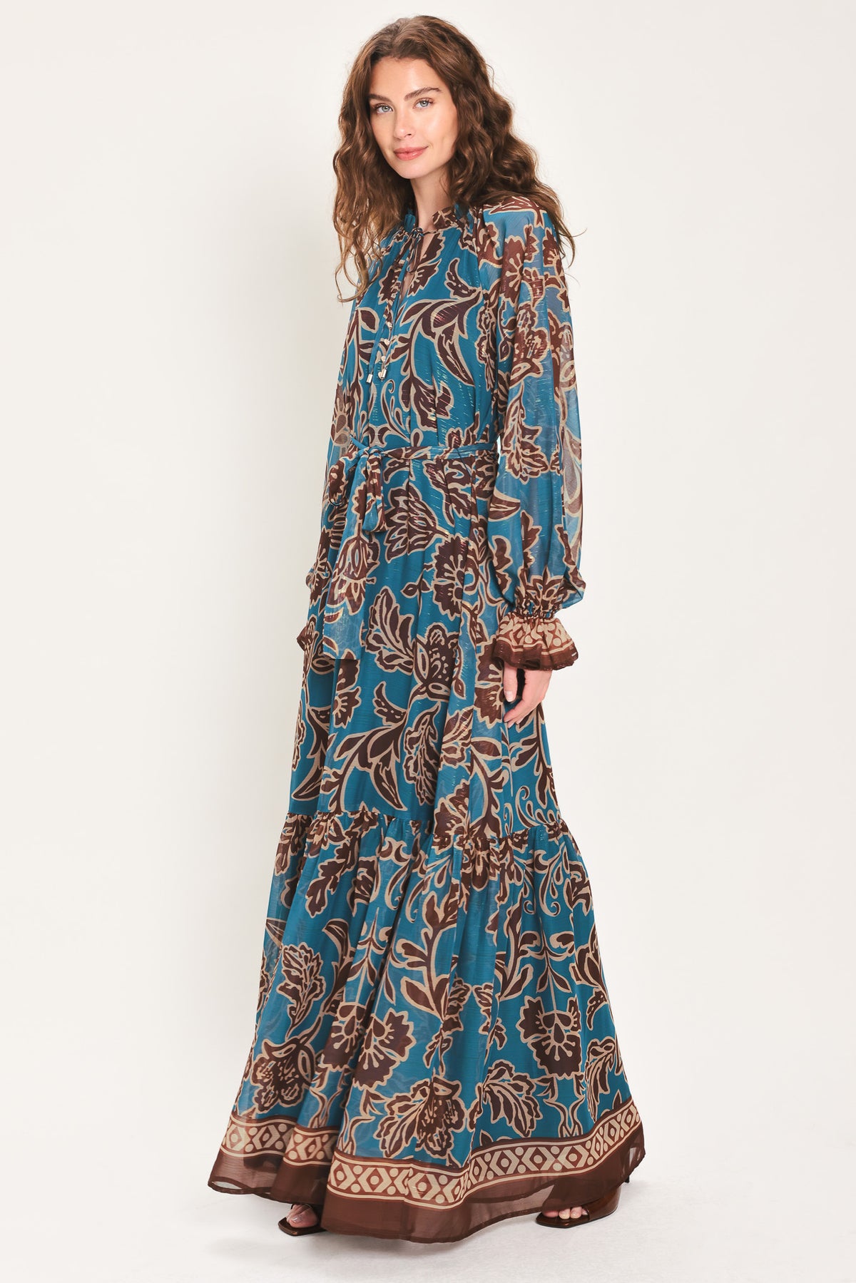 Woman wearing a teal blue long sleeve maxi dress with ruffled sleeves, tiered skirts, and brown floral print and neck tie | Flying Tomato