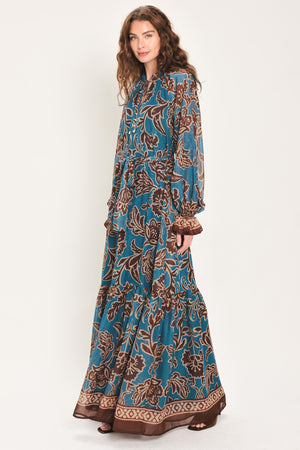 Woman wearing a teal blue long sleeve maxi dress with ruffled sleeves, tiered skirts, and brown floral print and neck tie | Flying Tomato