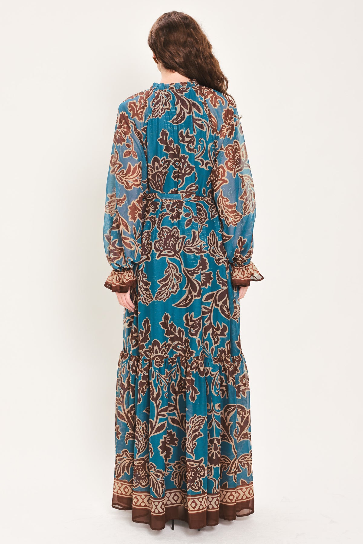 Woman wearing a teal blue long sleeve maxi dress with ruffled sleeves, tiered skirts, and brown floral print and neck tie | Flying Tomato
