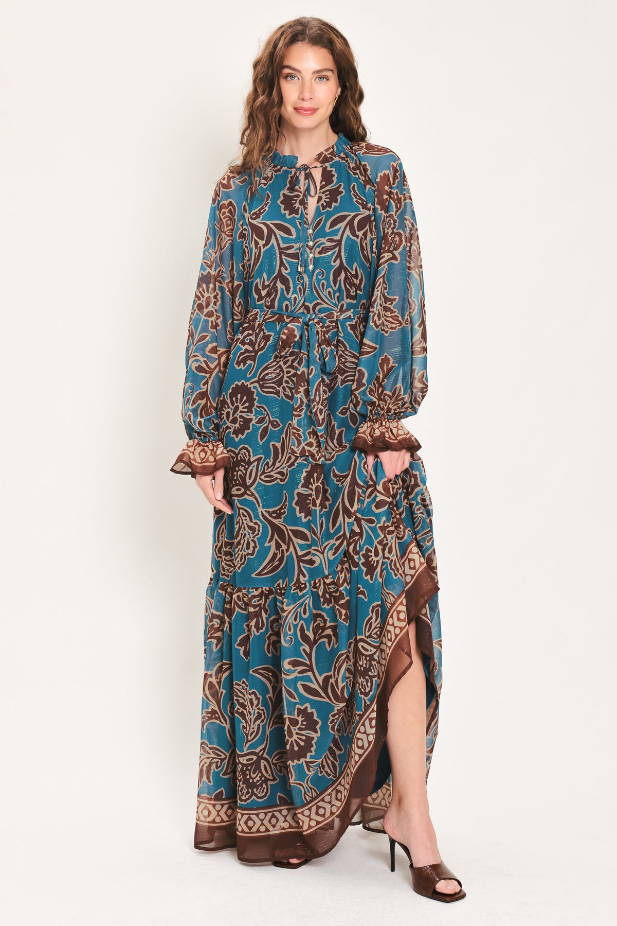Woman wearing a teal blue long sleeve maxi dress with ruffled sleeves, tiered skirts, and brown floral print and neck tie | Flying Tomato