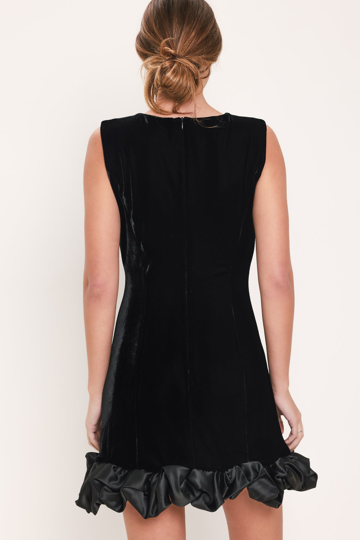 Woman wearing a sleeveless round neck black velvet mini dress with a satin ruffle at the hemline | Flying Tomato
