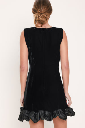 Woman wearing a sleeveless round neck black velvet mini dress with a satin ruffle at the hemline | Flying Tomato