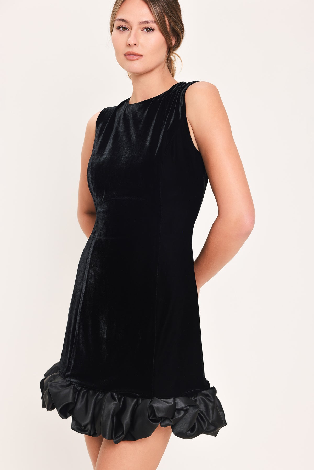 Woman wearing a sleeveless round neck black velvet mini dress with a satin ruffle at the hemline | Flying Tomato