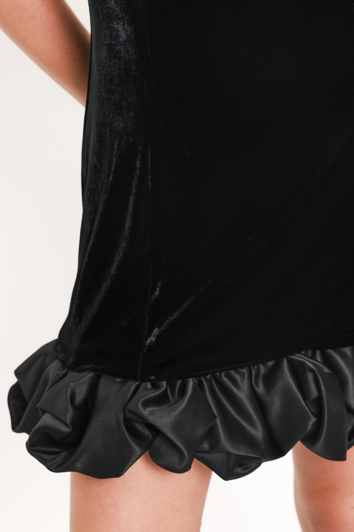 Woman wearing a sleeveless round neck black velvet mini dress with a satin ruffle at the hemline | Flying Tomato