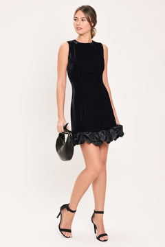 Woman wearing a sleeveless round neck black velvet mini dress with a satin ruffle at the hemline | Flying Tomato