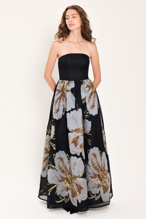 Woman wearing a strapless black maxi dress with a bluish grey floral print and gold details printed on the skirt | Flying Tomato