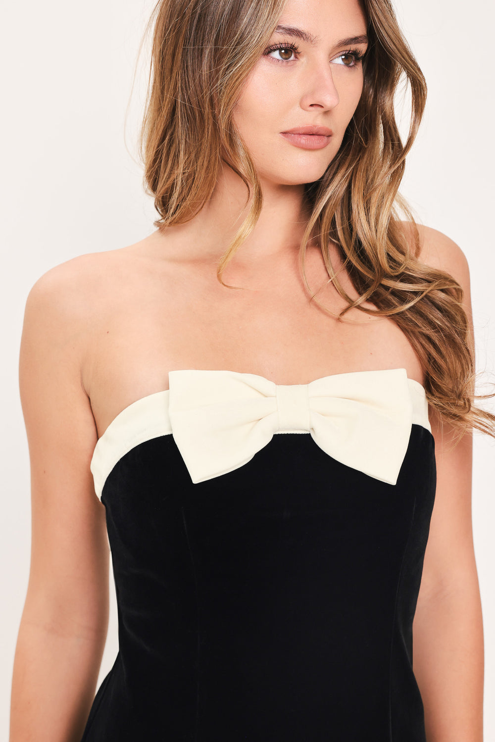 A woman modeling a strapless velvet black mini dress with a large cream bow at the bust | Flying Tomato