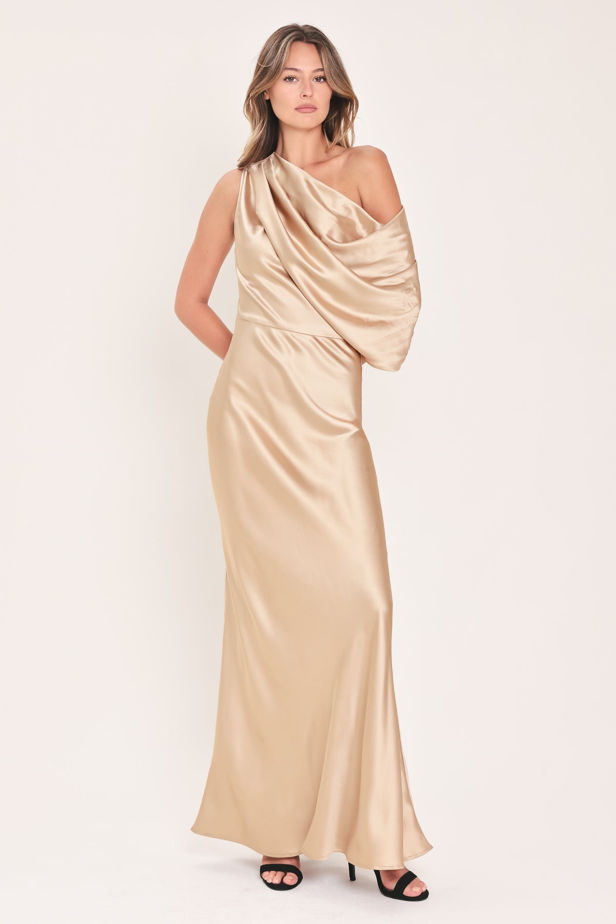 A woman modeling a floor-length draped satin gown in champagne beige, featuring an asymmetrical draped neckline and a flowing skirt | Flying Tomato