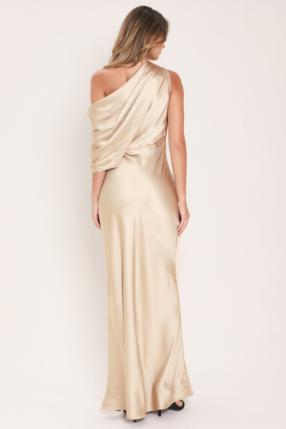 A woman modeling a floor-length, one-shoulder satin gown from the back, showing a draped neckline and a fitted skirt in a warm champagne beige color | Flying Tomato