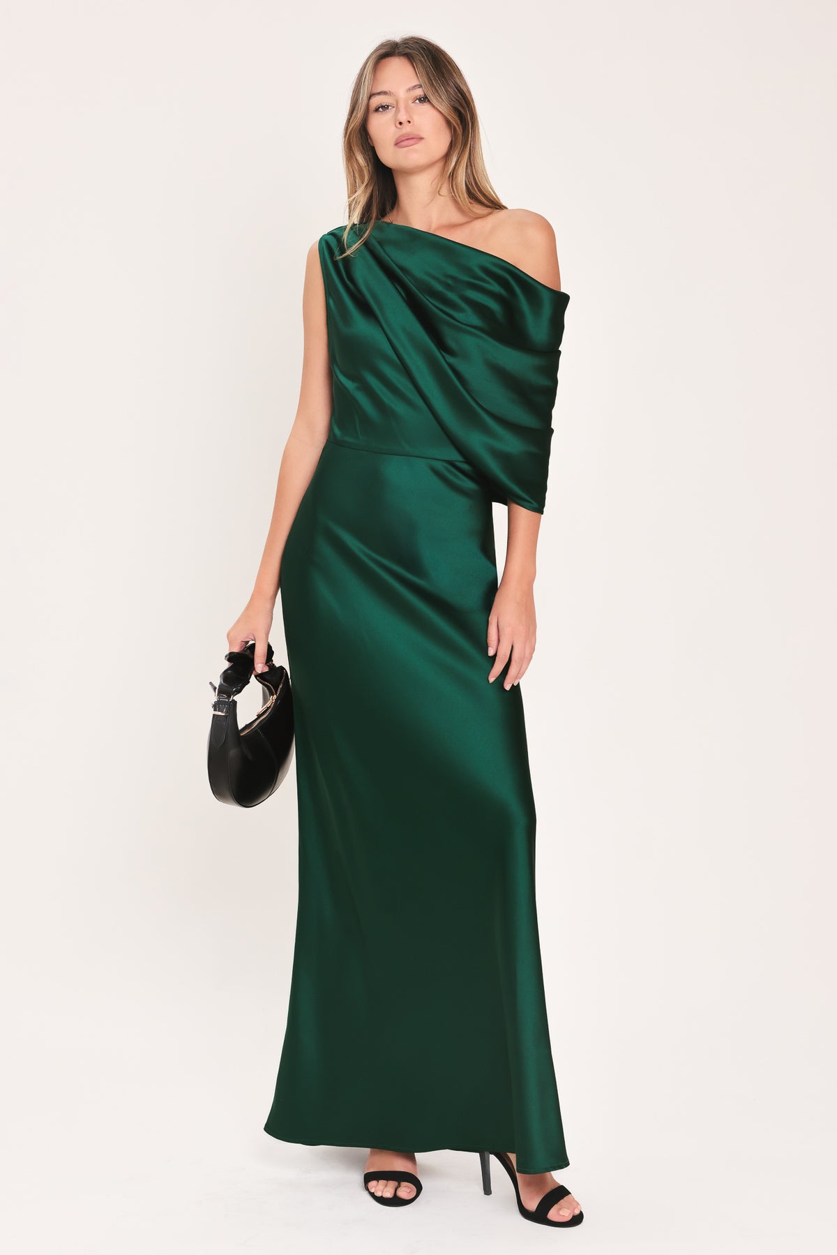 A woman modeling a floor-length maxi emerald green satin dress with draped asymmetrical detailing and a draped shoulder design | Flying Tomato