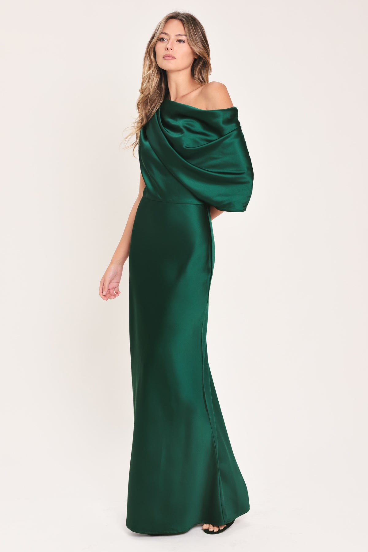 A woman modeling a floor-length maxi emerald green satin dress with draped asymmetrical detailing and a draped shoulder design | Flying Tomato