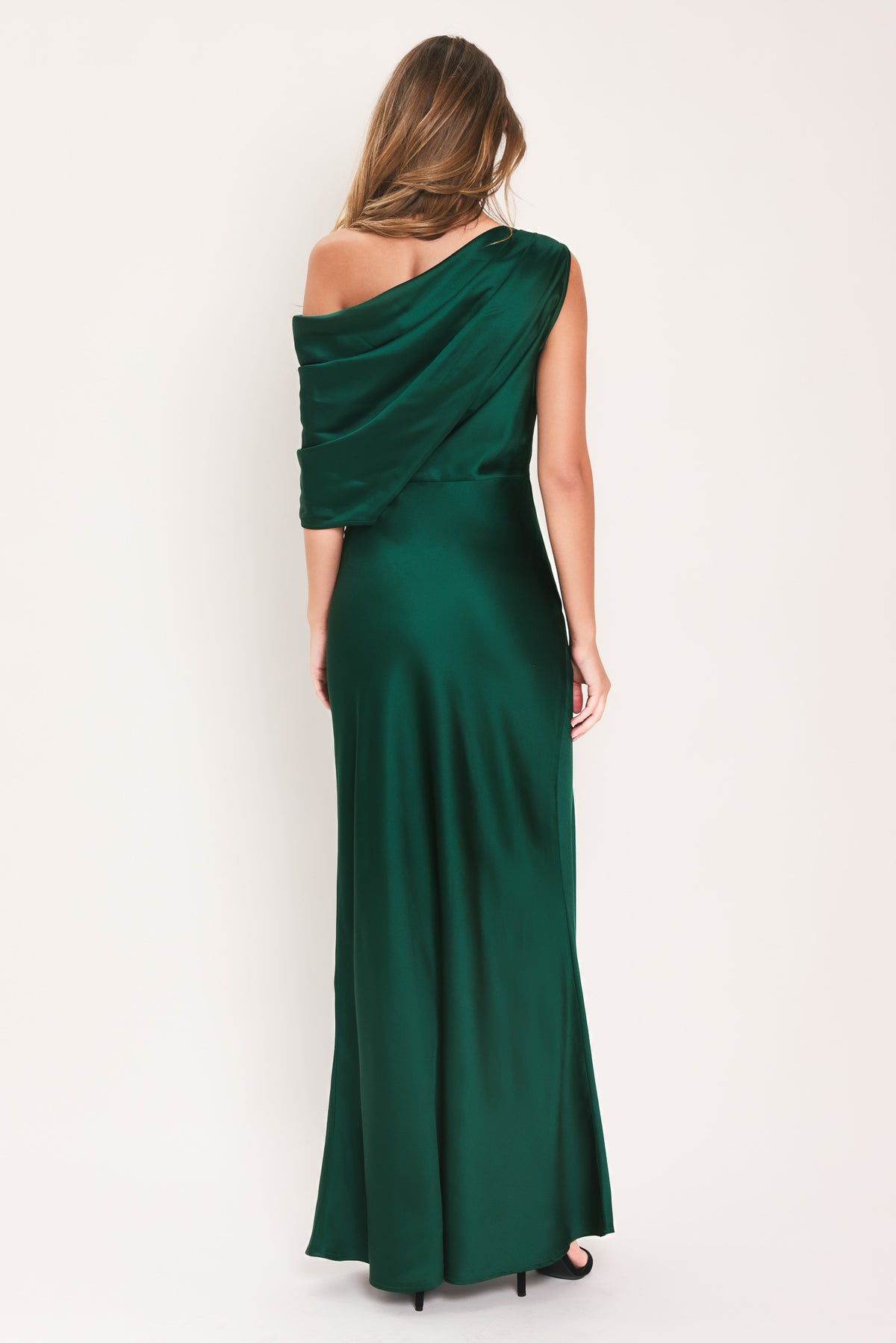 A woman modeling a floor-length maxi emerald green satin dress with draped asymmetrical detailing and a draped shoulder design | Flying Tomato