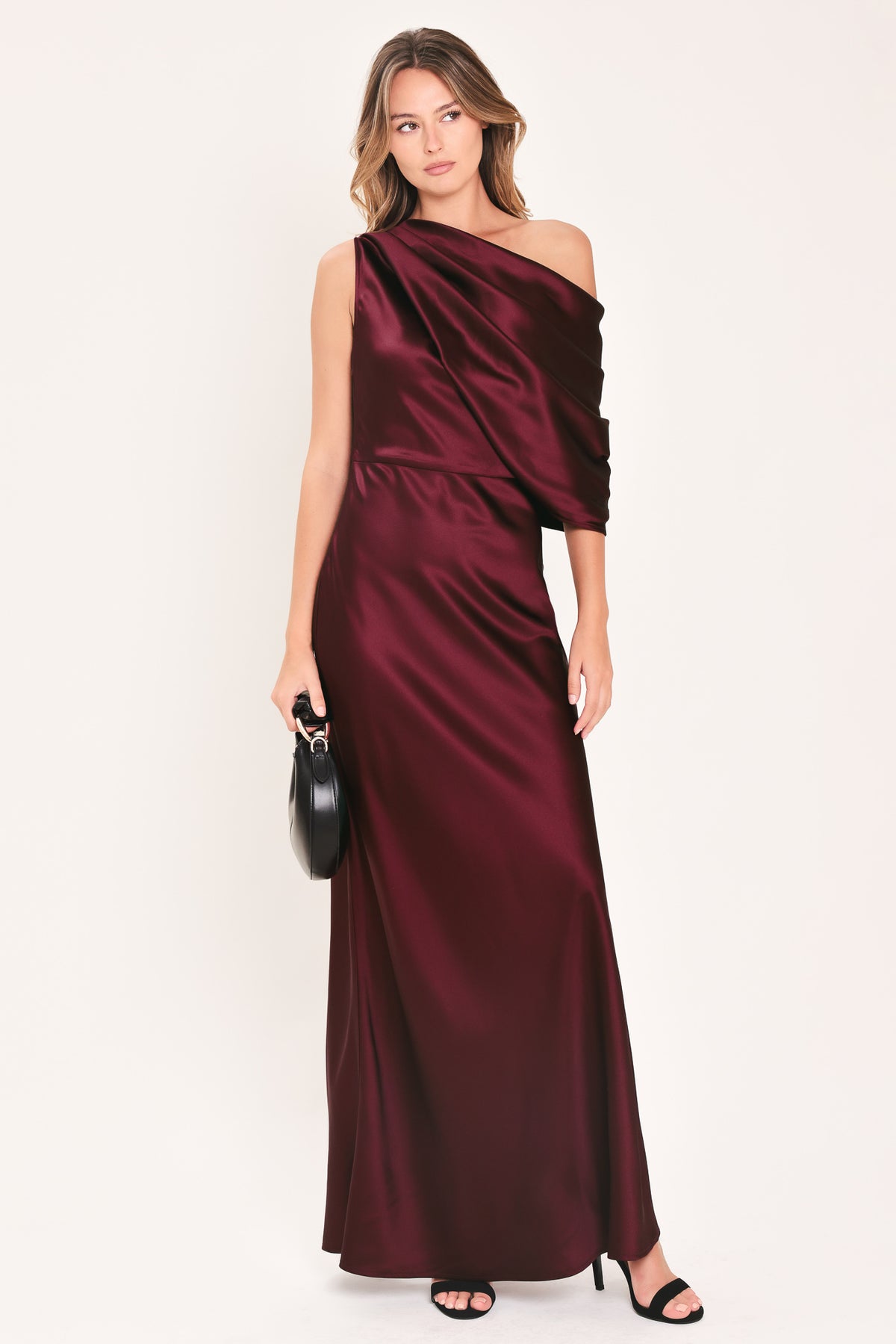 A woman modeling a floor-length maxi draped satin gown in deep burgundy wine, featuring an asymmetrical draped neckline and one sleeve | Flying Tomato