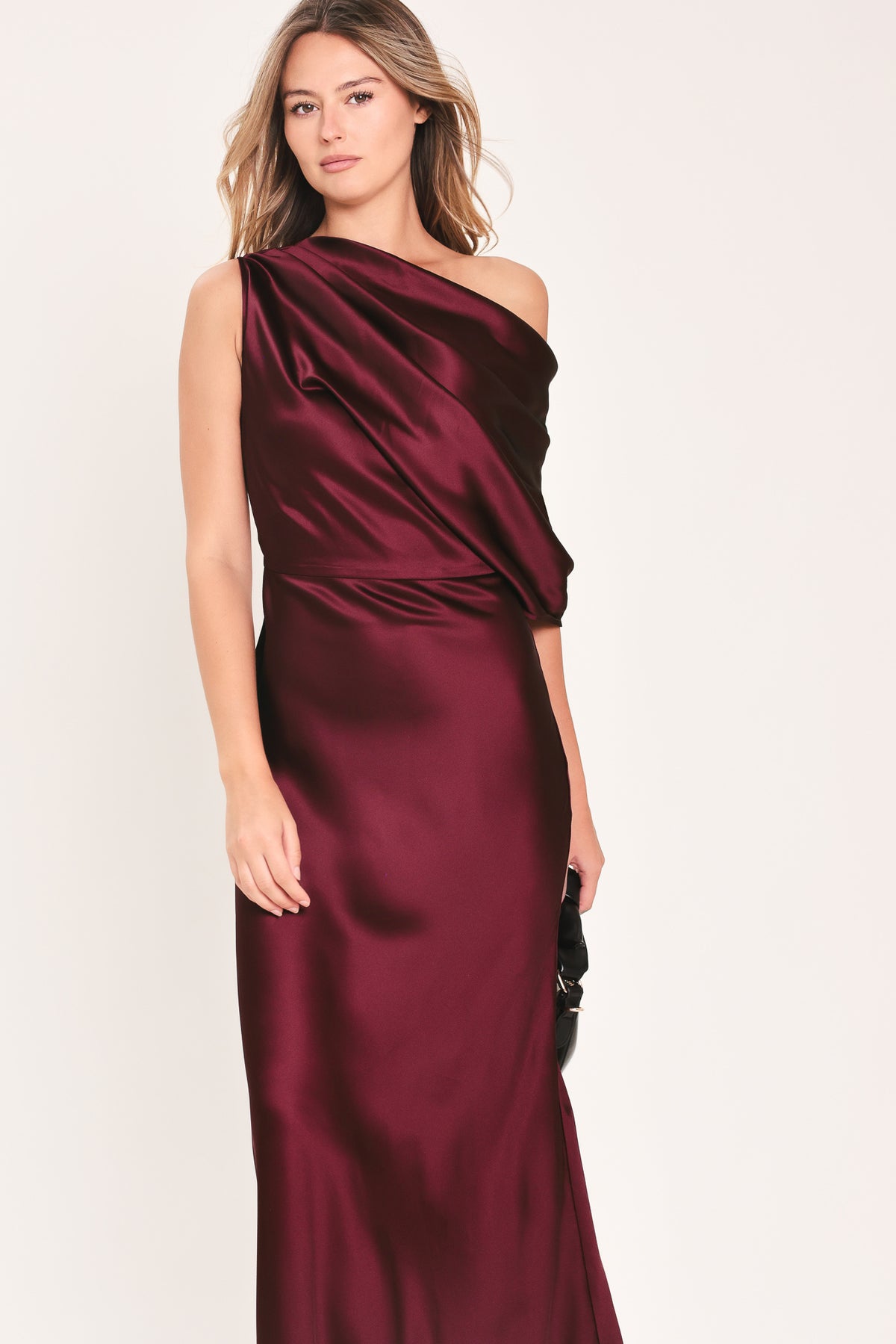 A woman modeling a floor-length maxi draped satin gown in deep burgundy wine, featuring an asymmetrical draped neckline and one sleeve | Flying Tomato