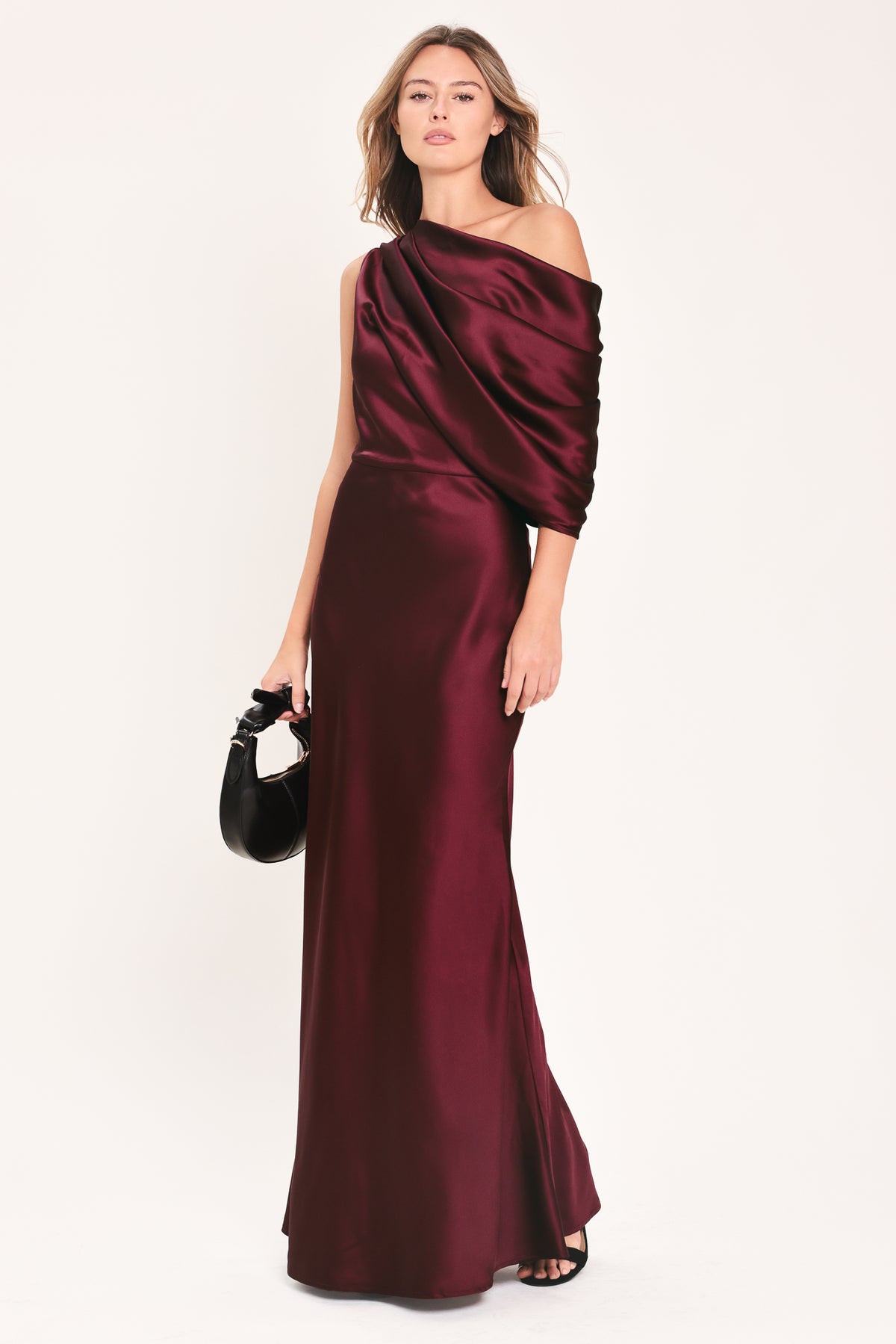 A woman modeling a floor-length maxi draped satin gown in deep burgundy wine, featuring an asymmetrical draped neckline and one sleeve | Flying Tomato