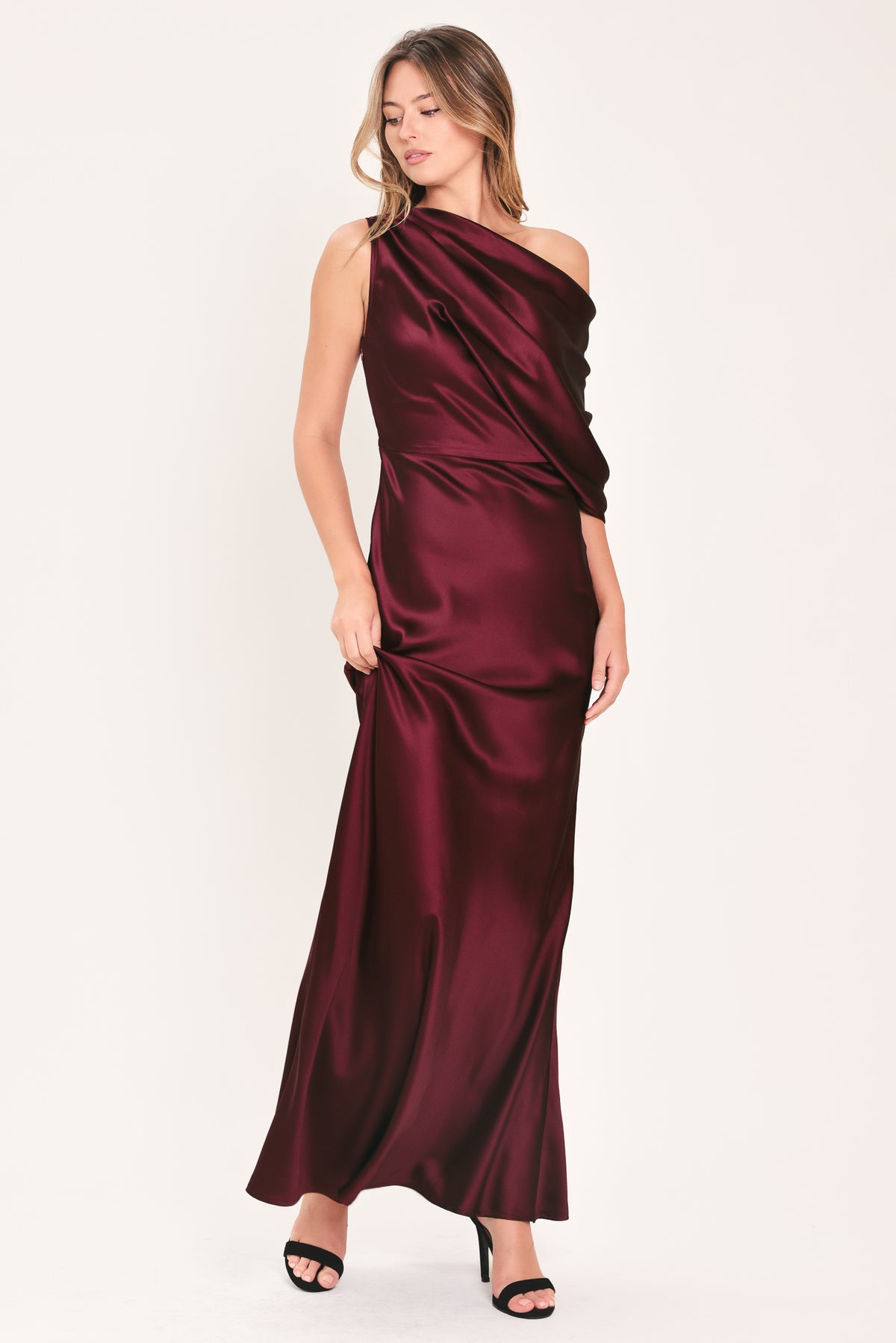 A woman modeling a floor-length maxi draped satin gown in deep burgundy wine, featuring an asymmetrical draped neckline and one sleeve | Flying Tomato