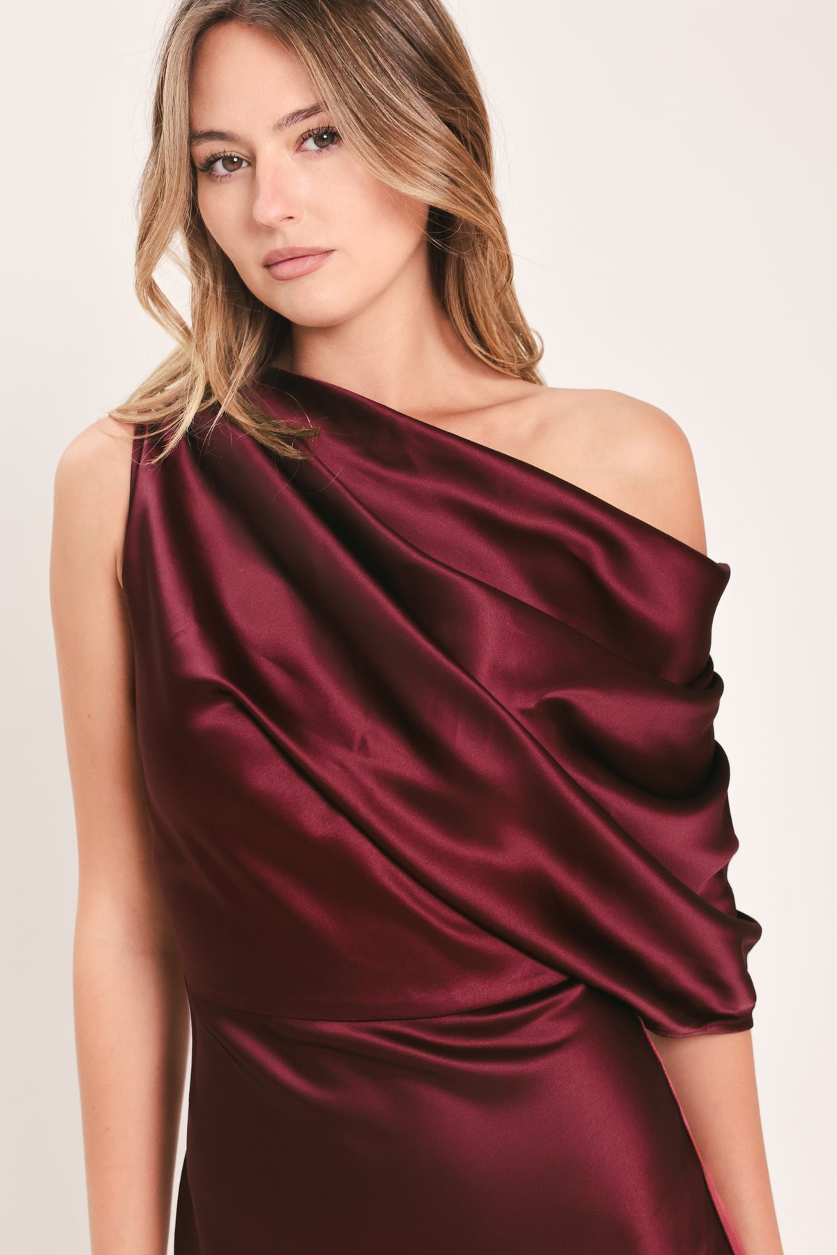 A woman modeling a floor-length maxi draped satin gown in deep burgundy wine, featuring an asymmetrical draped neckline and one sleeve | Flying Tomato