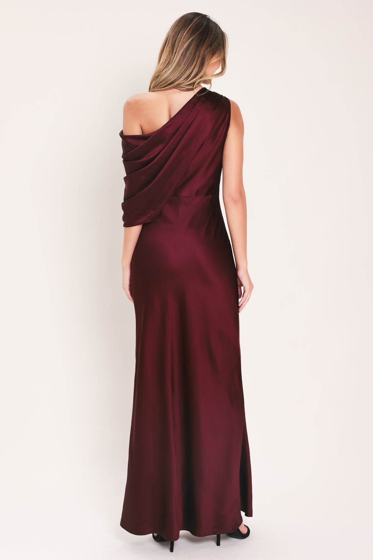 A woman modeling a floor-length maxi draped satin gown in deep burgundy wine, featuring an asymmetrical draped neckline and one sleeve | Flying Tomato