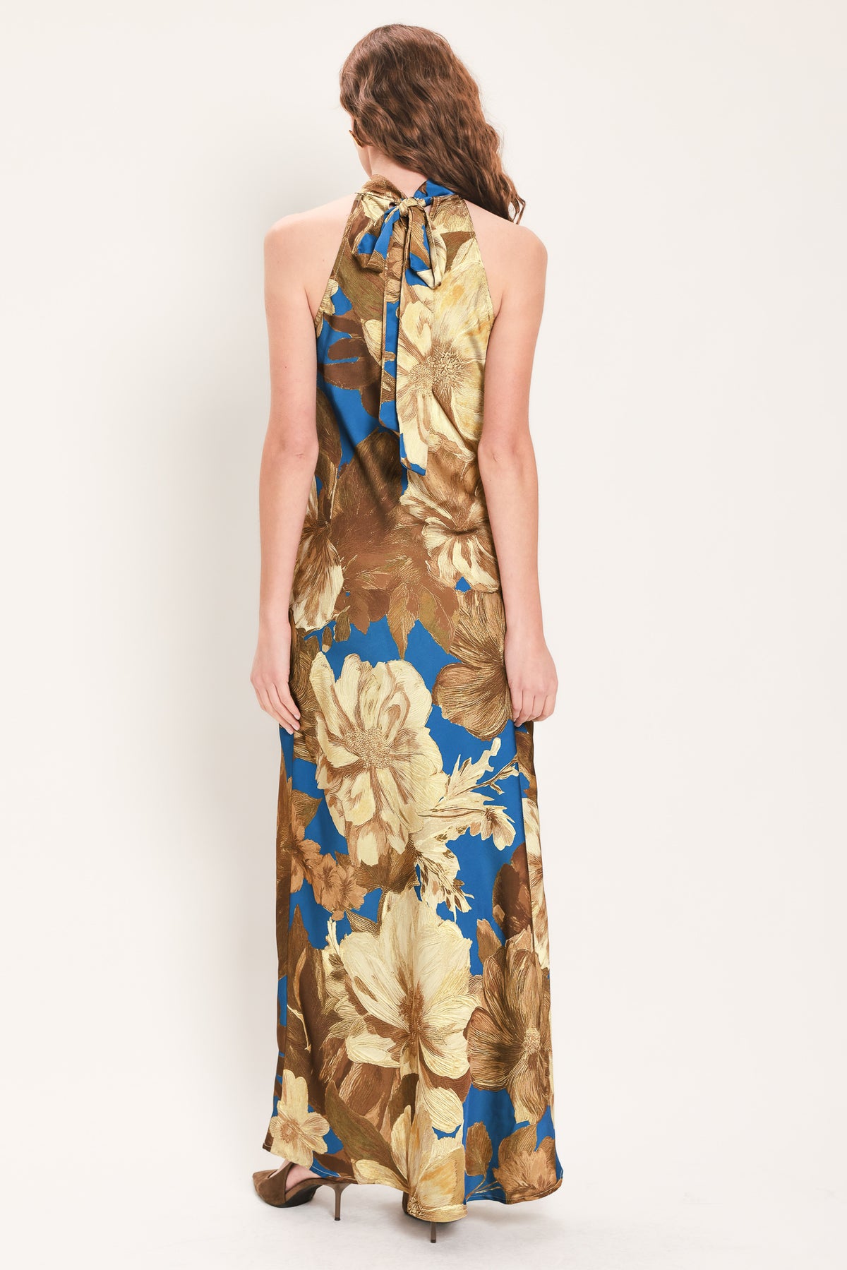 Woman wearing a sleeveless blue satin maxi dress with a mock halter neckline and gold and brown floral pattern | Flying Tomato