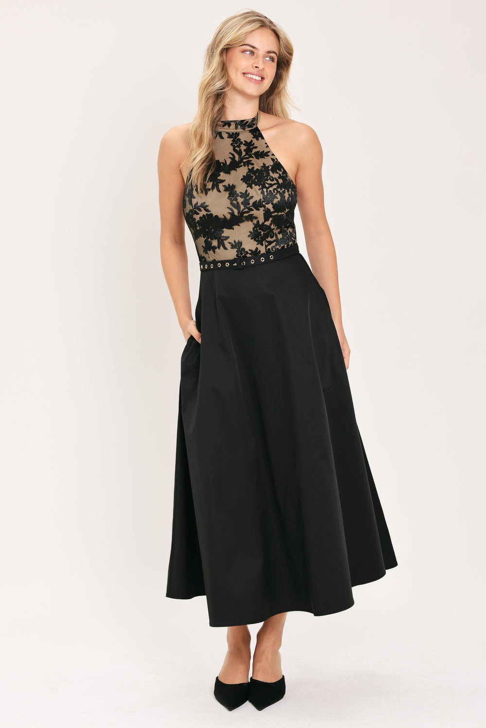 Woman wearing a halter mock neck midid dress with beige top base and black floral lace over and black skirt and black waist belt | Flying Tomato