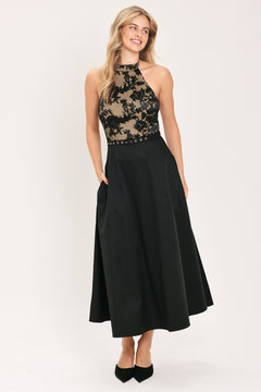 Woman wearing a halter mock neck midid dress with beige top base and black floral lace over and black skirt and black waist belt | Flying Tomato