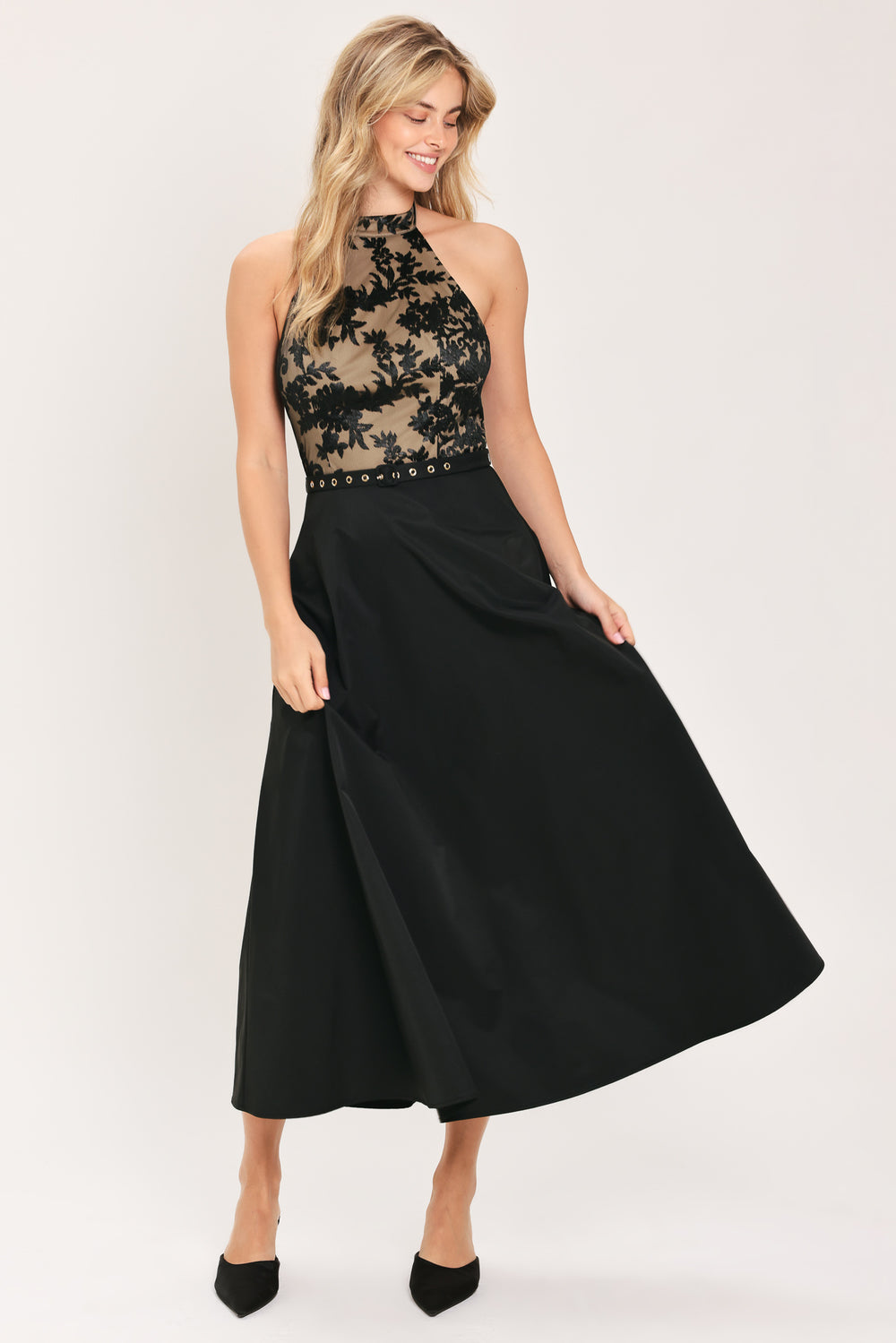 Woman wearing a halter mock neck midid dress with beige top base and black floral lace over and black skirt and black waist belt | Flying Tomato