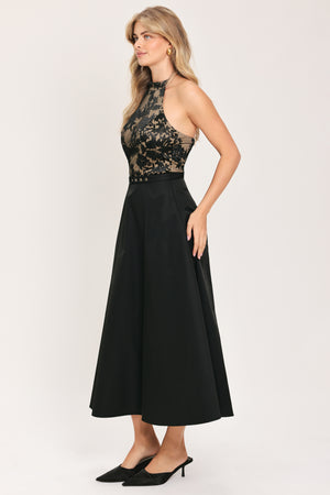 Woman wearing a halter mock neck midid dress with beige top base and black floral lace over and black skirt and black waist belt | Flying Tomato