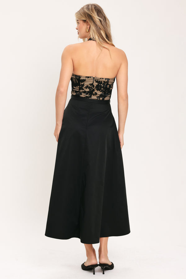 Woman wearing a halter mock neck midid dress with beige top base and black floral lace over and black skirt and black waist belt | Flying Tomato