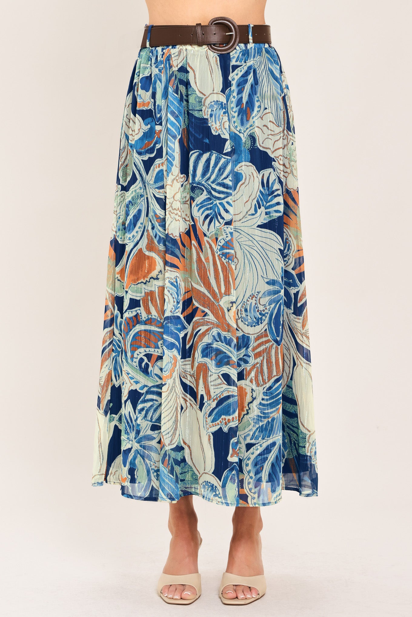 A woman wearing a high-waisted, midi-length maxi skirt featuring a vibrant tropical leaf print in shades of blue, orange, and cream. The skirt also has a brown leather belt | Flying Tomato