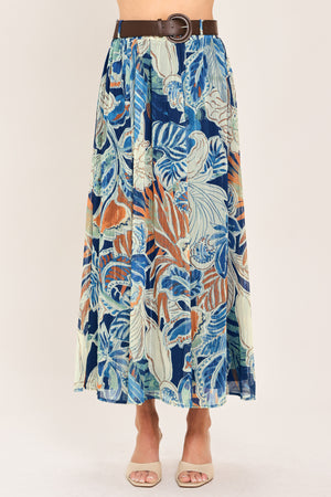 A woman wearing a high-waisted, midi-length maxi skirt featuring a vibrant tropical leaf print in shades of blue, orange, and cream. The skirt also has a brown leather belt | Flying Tomato