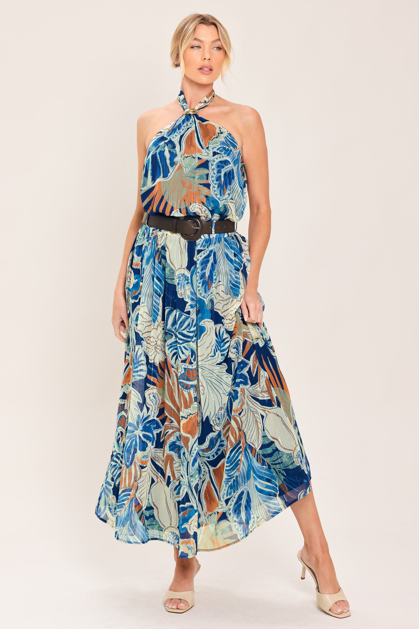 A woman wearing a high-waisted, midi-length maxi skirt featuring a vibrant tropical leaf print in shades of blue, orange, and cream. The skirt also has a brown leather belt | Flying Tomato