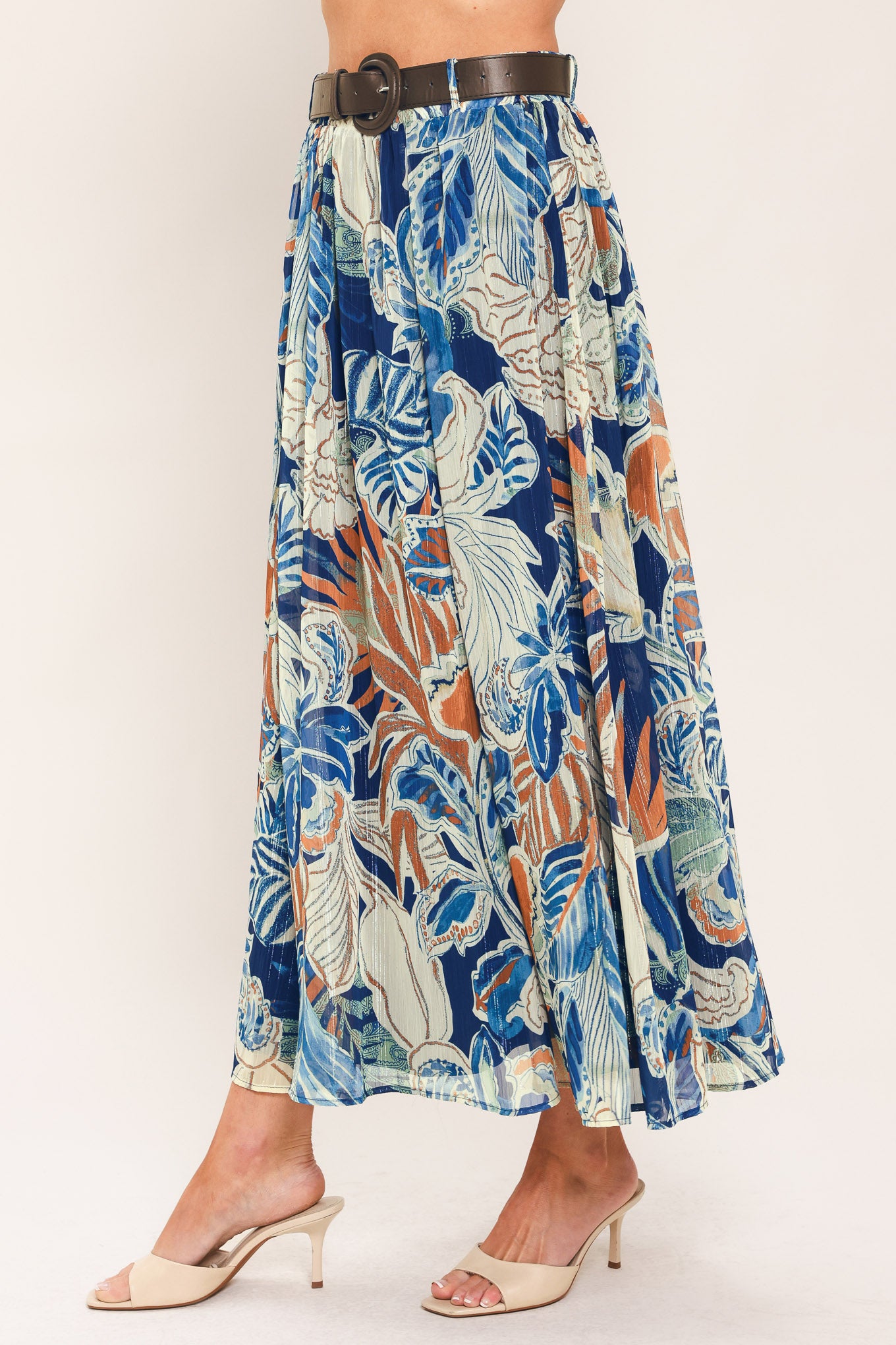A woman wearing a high-waisted, midi-length maxi skirt featuring a vibrant tropical leaf print in shades of blue, orange, and cream. The skirt also has a brown leather belt | Flying Tomato