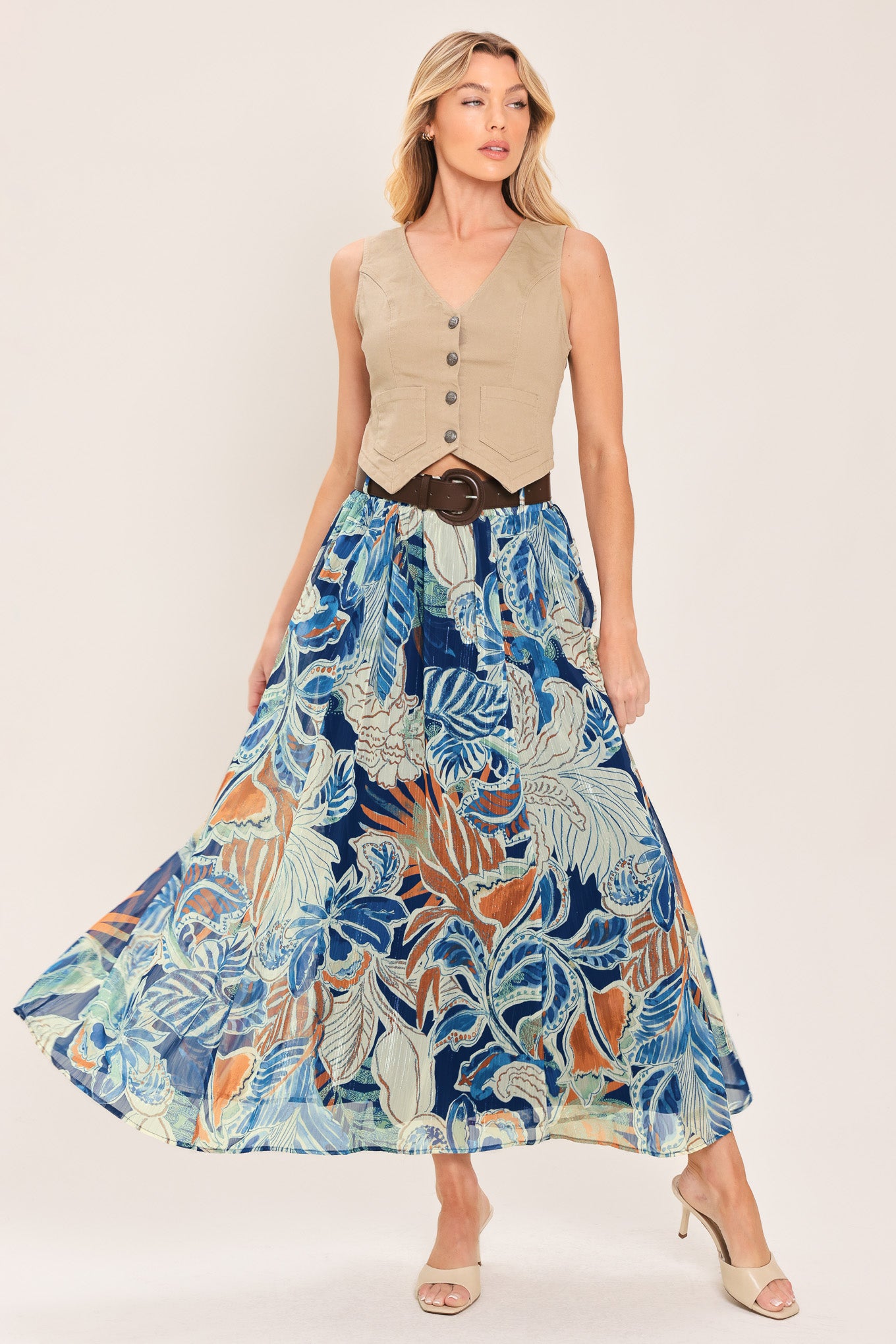 A woman wearing a high-waisted, midi-length maxi skirt featuring a vibrant tropical leaf print in shades of blue, orange, and cream. The skirt also has a brown leather belt | Flying Tomato