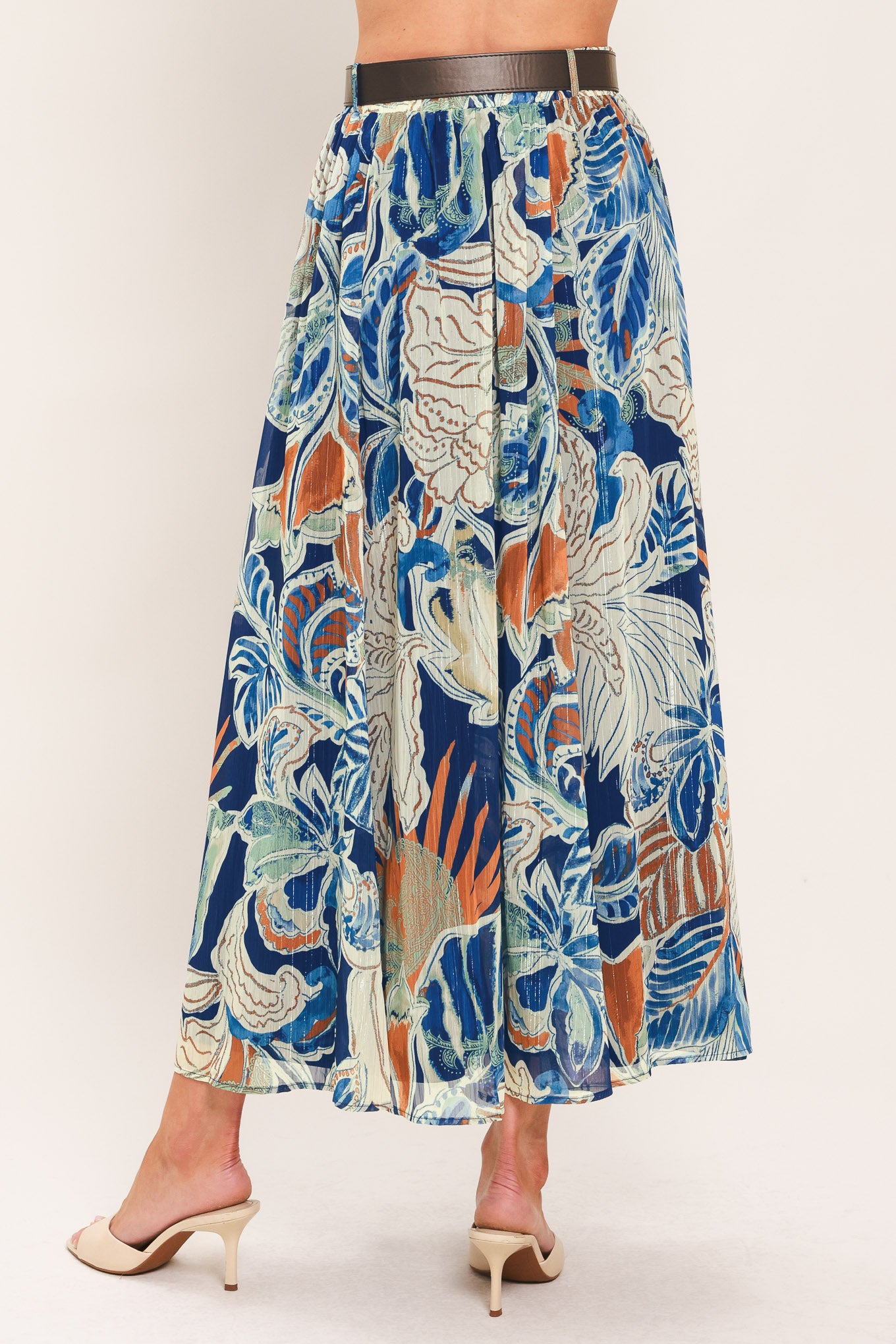 A woman wearing a high-waisted, midi-length maxi skirt featuring a vibrant tropical leaf print in shades of blue, orange, and cream. The skirt also has a brown leather belt | Flying Tomato