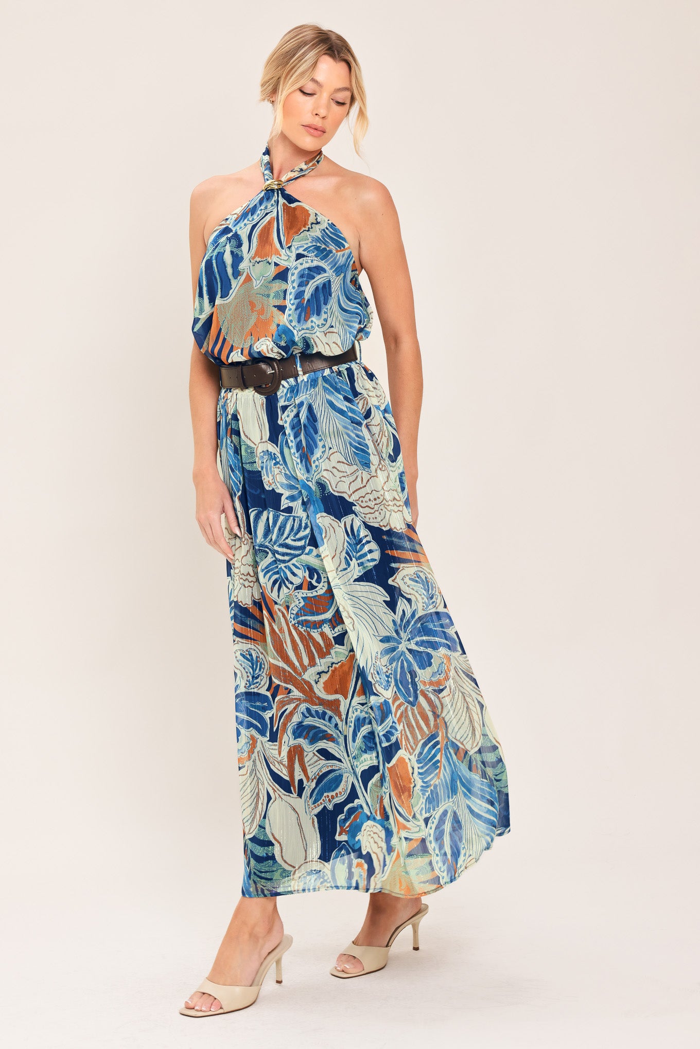 A woman wearing a high-waisted, midi-length maxi skirt featuring a vibrant tropical leaf print in shades of blue, orange, and cream. The skirt also has a brown leather belt | Flying Tomato