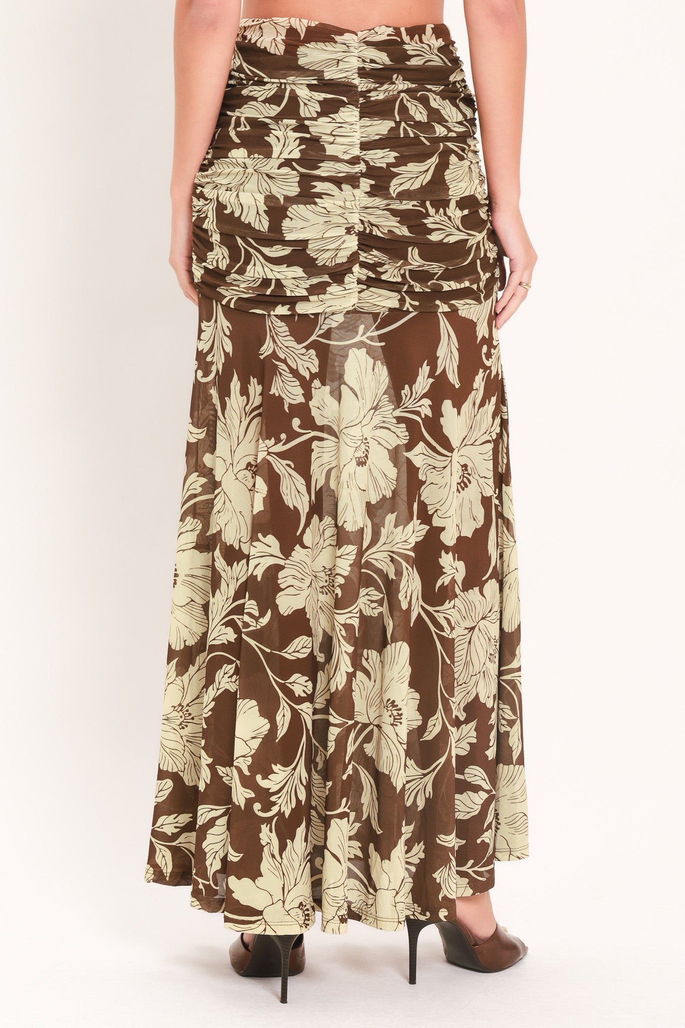 A woman wearing a brown and beige floral print maxi skirt with a ruched waistband, paired with dark brown high heels, shown from the back against a plain white studio background.