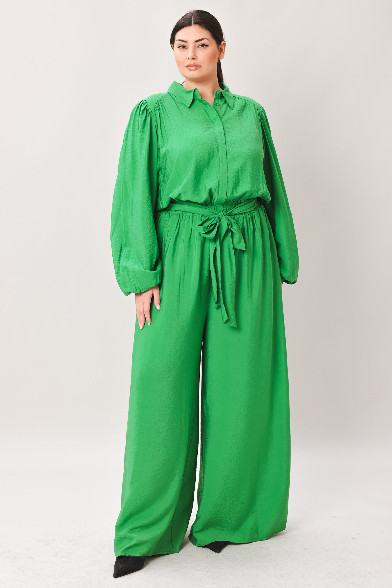Woman wearing a green jumpsuit featuring shirt collar, button down, long sleeve, elasticized waist, self sash tie, side pockets and wide leg on a white background | Flying Tomato