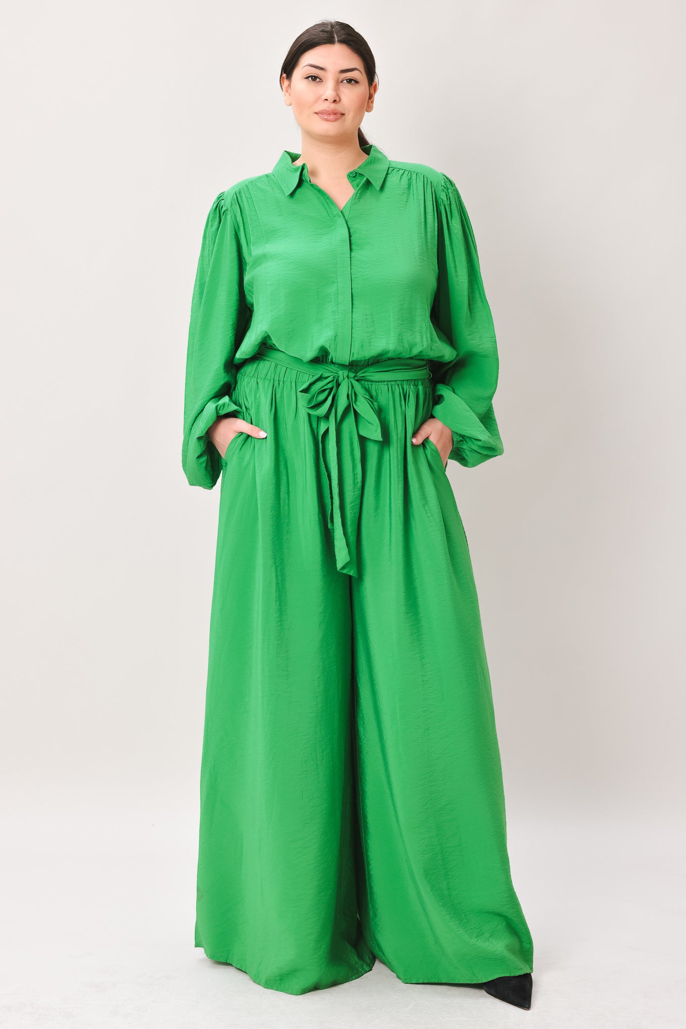 Woman wearing a green jumpsuit featuring shirt collar, button down, long sleeve, elasticized waist, self sash tie, side pockets and wide leg on a white background | Flying Tomato