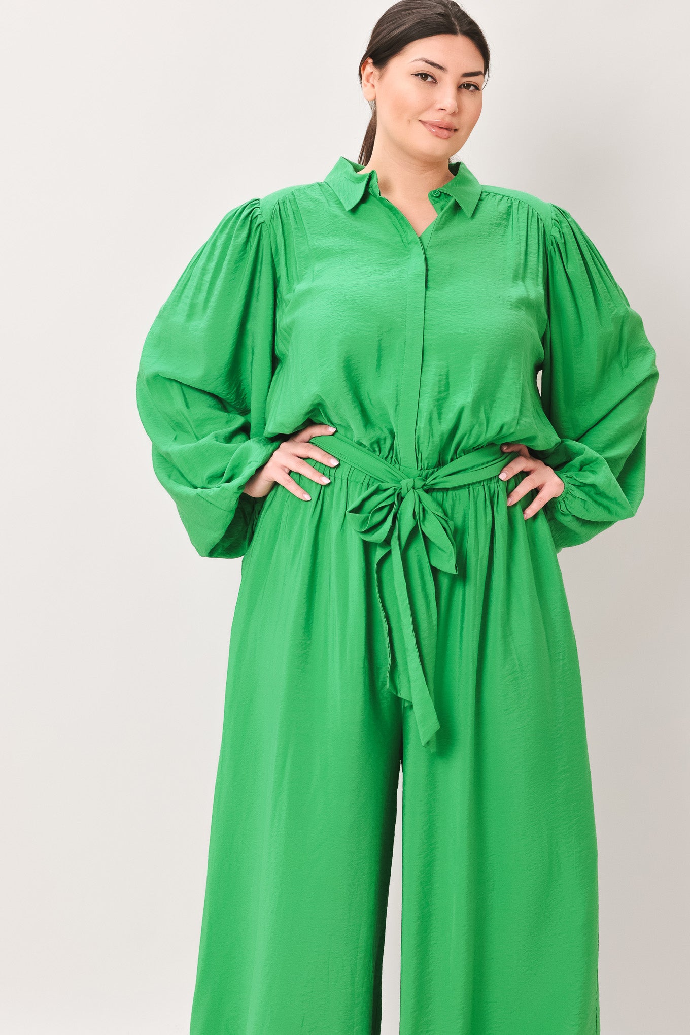 Woman wearing a green jumpsuit featuring shirt collar, button down, long sleeve, elasticized waist, self sash tie, side pockets and wide leg on a white background | Flying Tomato