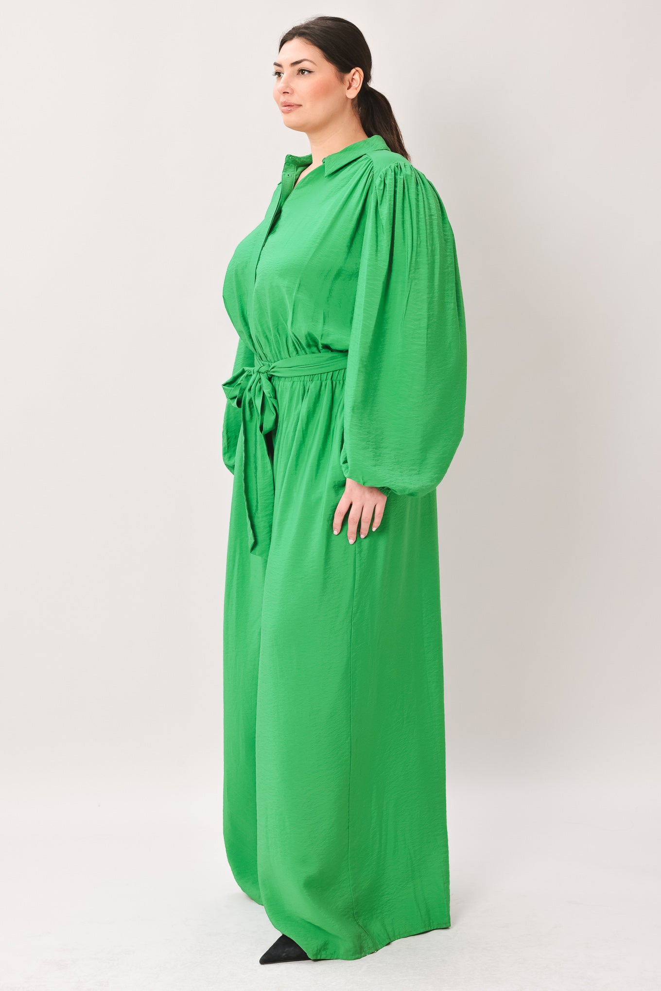 Woman wearing a green jumpsuit featuring shirt collar, button down, long sleeve, elasticized waist, self sash tie, side pockets and wide leg on a white background | Flying Tomato
