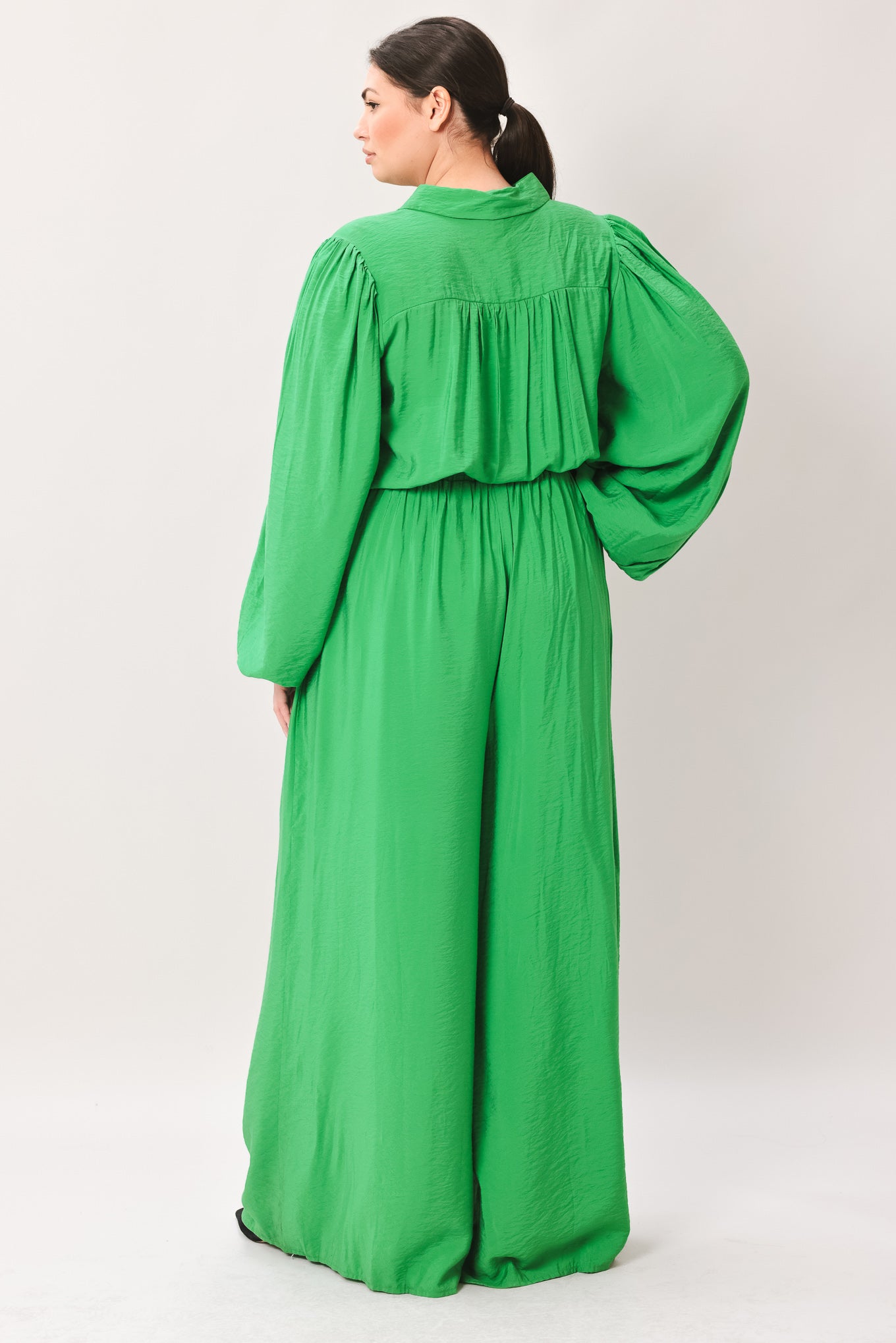 Woman wearing a green jumpsuit featuring shirt collar, button down, long sleeve, elasticized waist, self sash tie, side pockets and wide leg on a white background | Flying Tomato