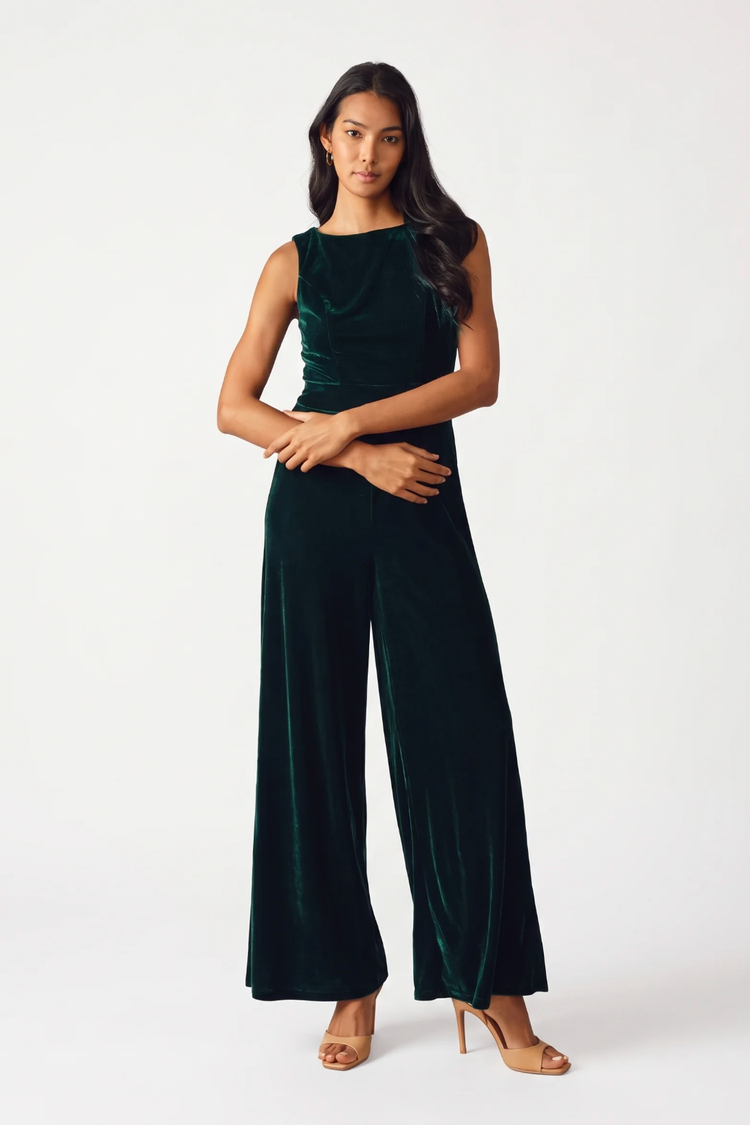 A woman modeling a dark green velvet sleeveless jumpsuit with wide legs, paired with nude high-heeled sandals, against a plain white studio background.