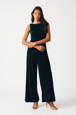 A woman modeling a dark green velvet sleeveless jumpsuit with wide legs, paired with nude high-heeled sandals, against a plain white studio background.