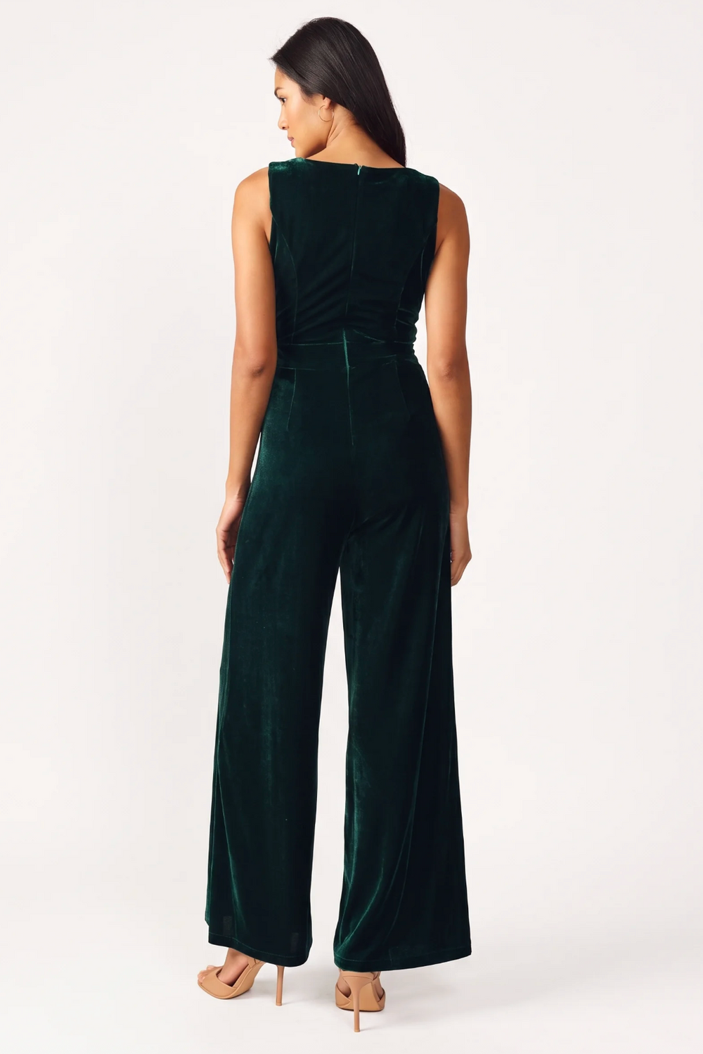 A woman modeling a dark green velvet sleeveless jumpsuit with wide legs, paired with nude high-heeled sandals, against a plain white studio background.
