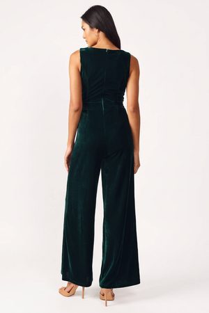 A woman viewed from the back wearing a sleeveless dark green velvet jumpsuit with wide legs, paired with nude high heels, against a plain white studio background.