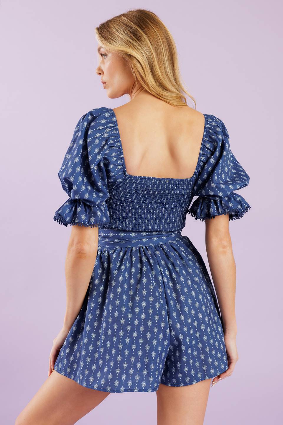 A textured woven romper featuring sweetheart neckline, short sleeve with lace trimmed ruffled cuff, smocked bodice, self sash belt and lace trimmed hemline.