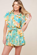 A printed woven romper featuring collar, button down, short cuffed sleeve, elasticized waist and self sash tie.