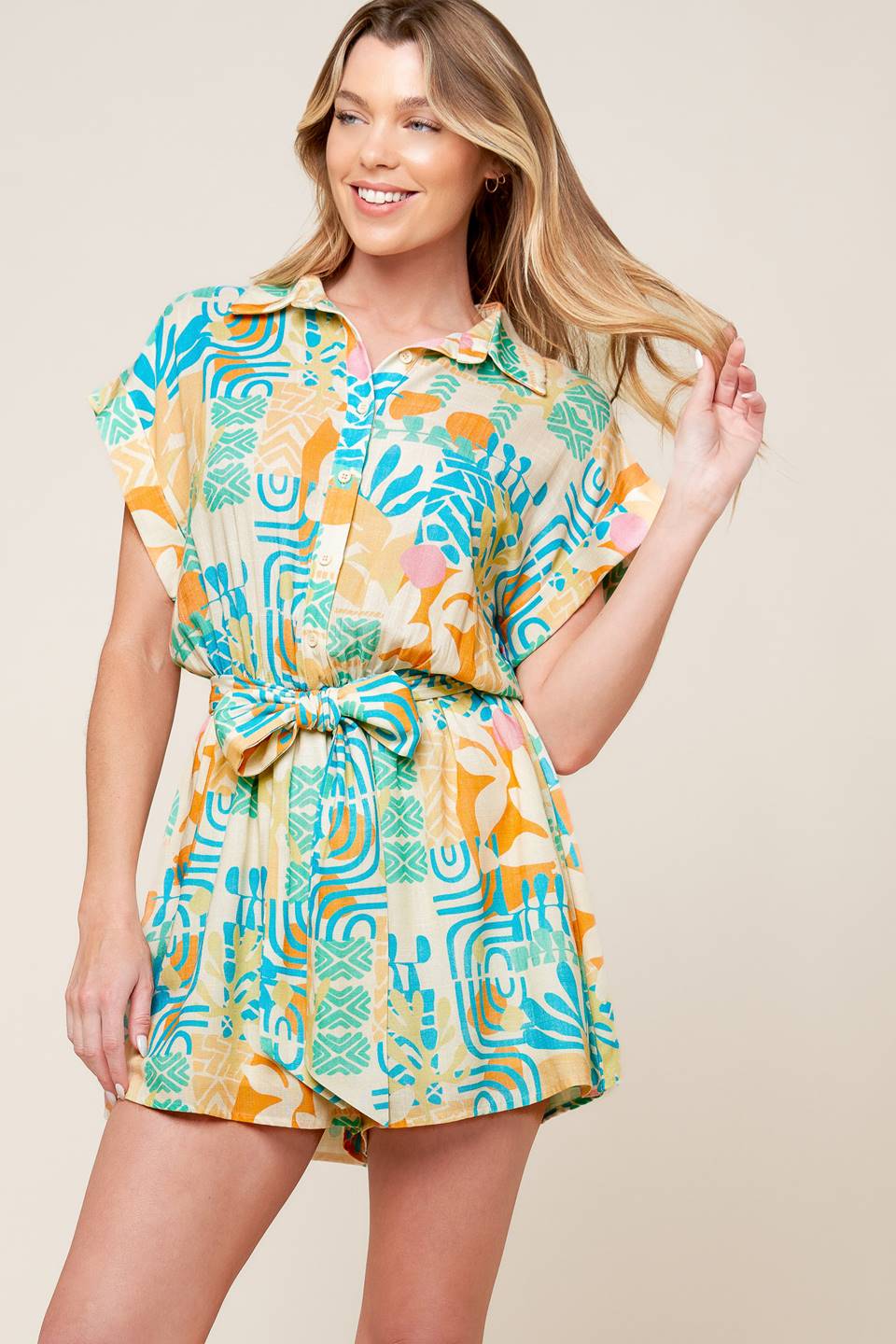 A printed woven romper featuring collar, button down, short cuffed sleeve, elasticized waist and self sash tie.